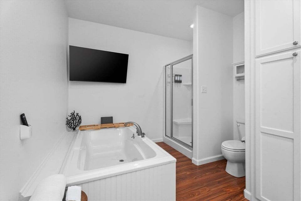Spacious primary bathroom with deep soaking tub, separate glass-enclosed shower, toilet area, wall-mounted TV and bright neutral finishes.