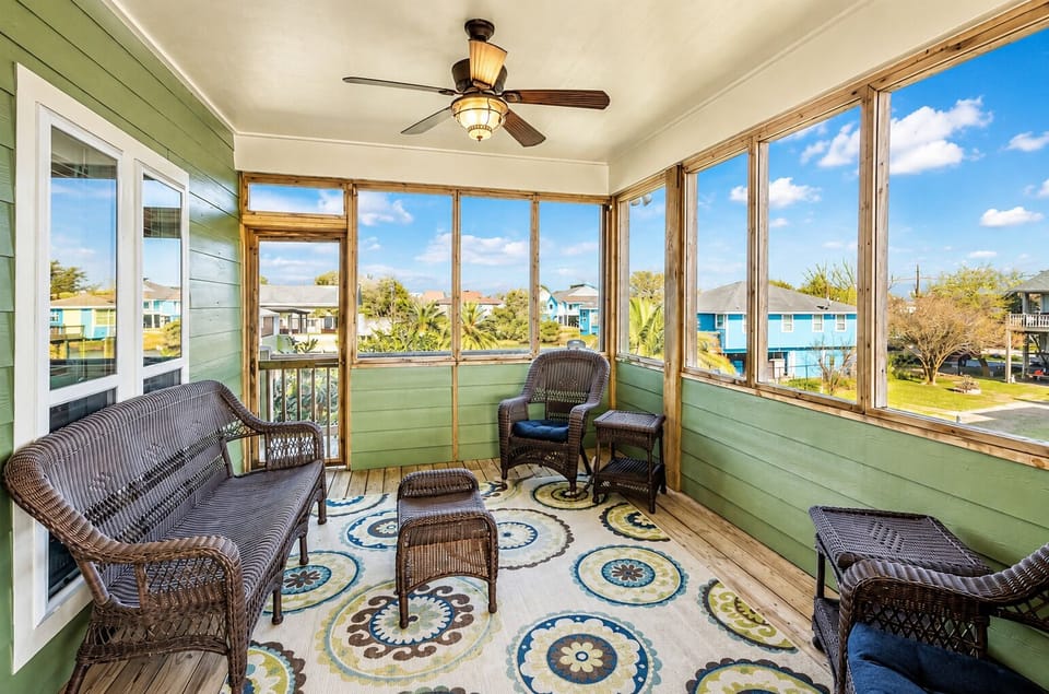Bright screened porch with wicker seating, ceiling fan, colorful area rug and wide neighborhood views, offering a comfortable indoor-outdoor lounge space.