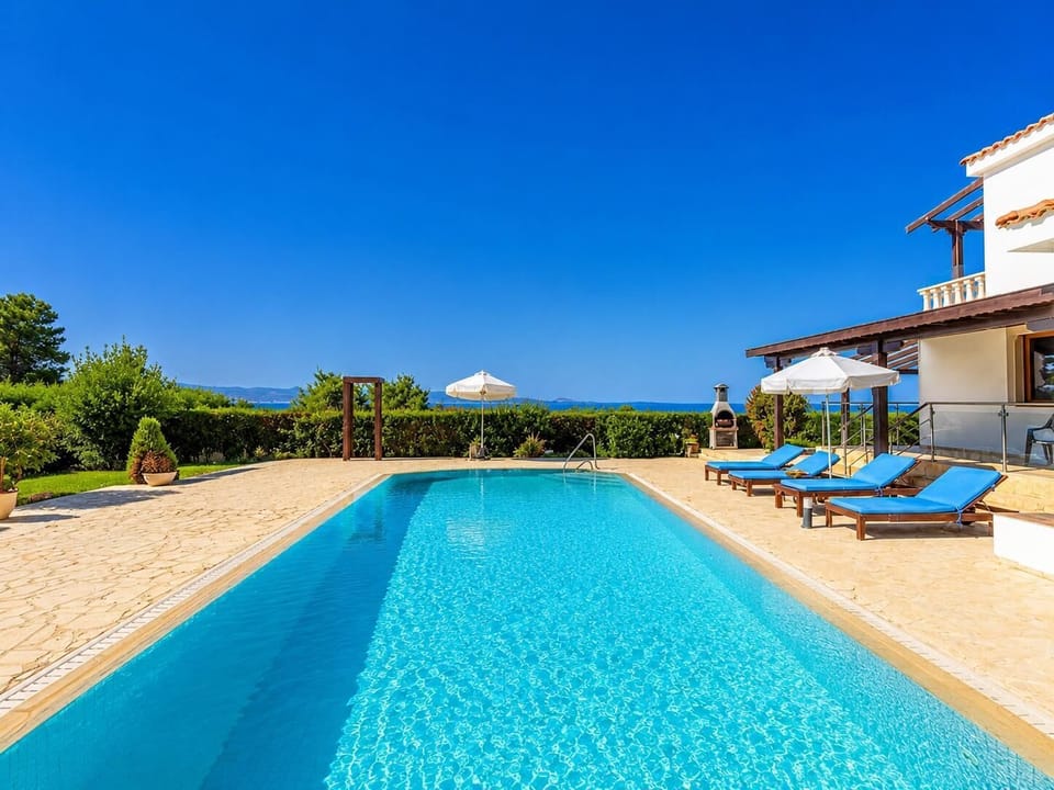 A luxurious villa pool terrace with blue sun loungers, white umbrellas, and sweeping sea views under a clear sky.