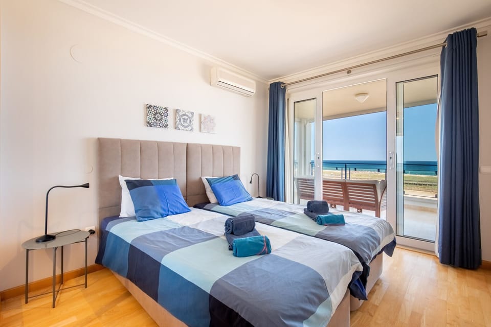 Welcome to your bright and inviting bedroom, designed for ultimate comfort and relaxation. You'll find two cozy single beds, perfectly arranged with soft blue and white checkered bedding, complemented by plush blue pillows and fresh towels. The highlight of this room is the direct access to your private balcony through large sliding glass doors, where you can step out and breathe in the fresh sea air while enjoying stunning, expansive views of the ocean and beach. This room provides a truly comfortable and refreshing coastal escape, complete with elegant wooden flooring and air conditioning for your comfort.