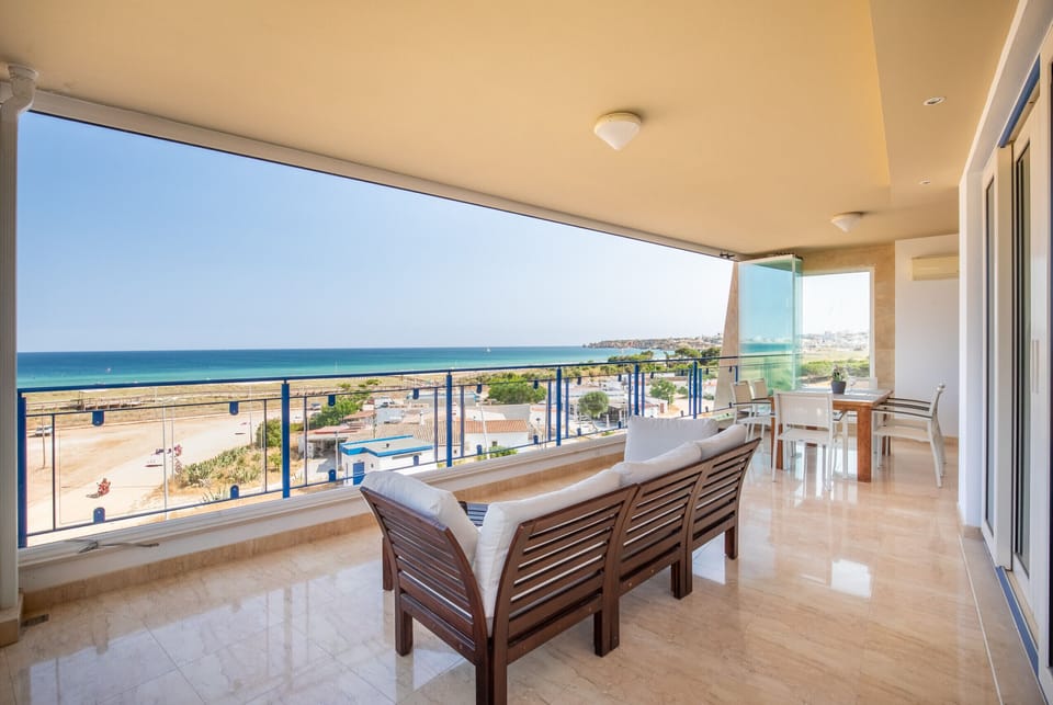 This spacious balcony offers breathtaking panoramic views of the ocean and the coastline, extending to the horizon. Furnished with comfortable wooden outdoor sofas with white cushions and a dining table with chairs, it's an ideal spot for guests to relax, dine with a view, and enjoy the refreshing sea breeze. The transparent glass railings ensure an unobstructed vista, allowing guests to fully appreciate the stunning seaside surroundings and identify the distinct coastal features.