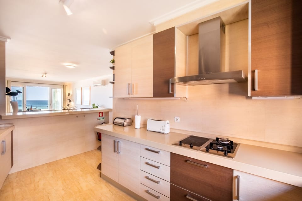 A functional and well-organized kitchen, integrated into the social space of the apartment. It has a spacious work area, modern cabinets, and essential appliances for everyday use. The counter serves as a support point for light meals or for socializing while cooking, always maintaining a visual connection with the living room and balcony with sea views.