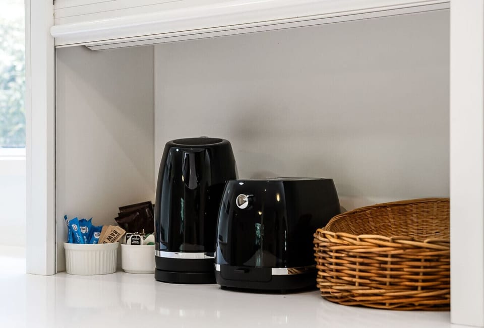 A kitchen corner with two black electric kettles placed on a white surface. Next to the kettles, there is a white ramekin containing tea packets and a wicker basket, suggesting a setup for making hot beverages.