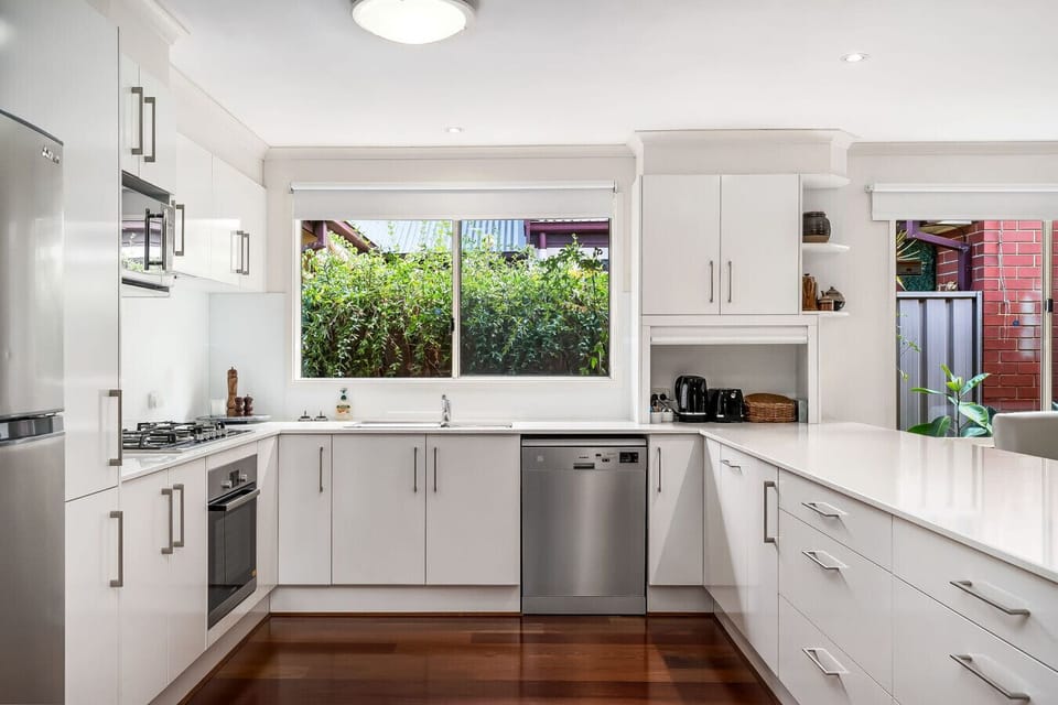 A modern, bright kitchen with white cabinetry and stainless steel appliances, including a fridge, oven, and dishwasher. The kitchen has a sleek design with a wooden floor and a large window offering a view of greenery outside, creating a fresh and airy at