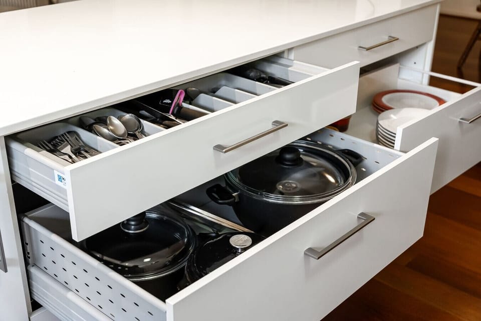 A kitchen cabinet with open drawers. The top drawer contains neatly organized cutlery such as forks, spoons, and knives, while the bottom drawer holds pots and pans. The cabinet has a sleek, white design with metal handles, and the background includes a w