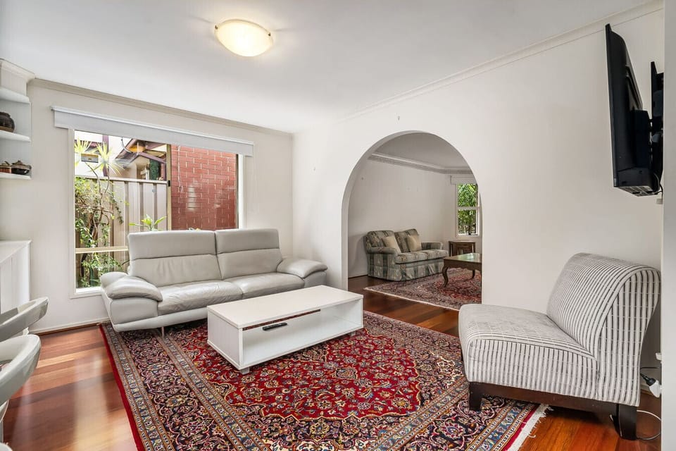 A cozy living room with a modern and clean design. It features a light-colored leather sofa, a striped armchair, and a decorative patterned rug on a wooden floor. There is an arched doorway leading to another room with a patterned sofa, and a flat-screen 