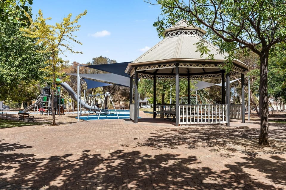 A park scene with a gazebo in the foreground, surrounded by trees and paved ground. In the background, there is a playground area with slides and other play equipment, set under a clear blue sky, creating a pleasant and inviting outdoor environment.
