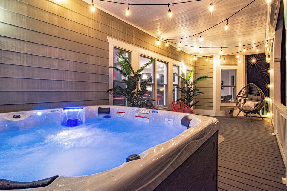 A cozy and inviting outdoor space featuring a bubbling hot tub illuminated with soft blue lighting, surrounded by warm string lights. The covered deck is adorned with lush greenery, stylish seating, and a relaxing egg chair, creating the perfect ambiance for unwinding.