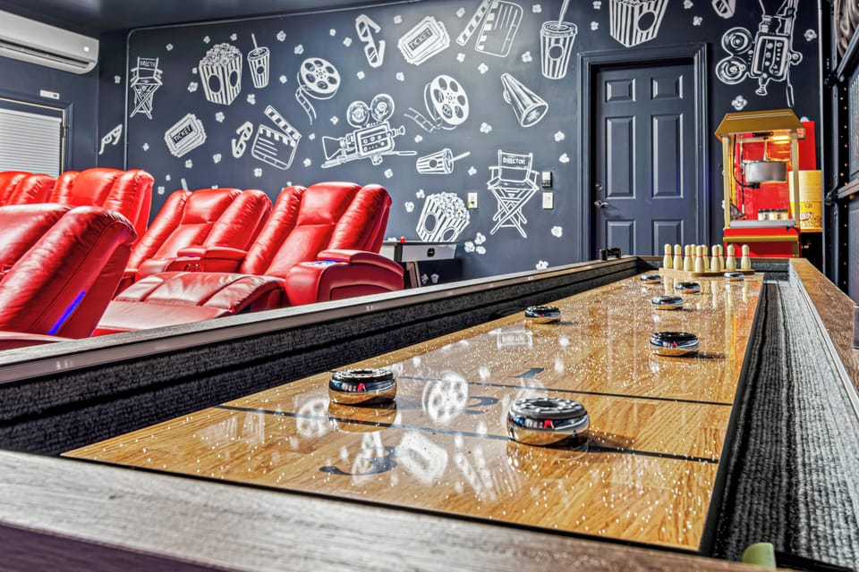 A stylish movie theater room featuring plush red reclining seats, a cinematic-themed mural, and a popcorn machine for the ultimate viewing experience. A sleek shuffleboard table in the foreground adds a fun touch, making this space perfect for both entertainment and relaxation.