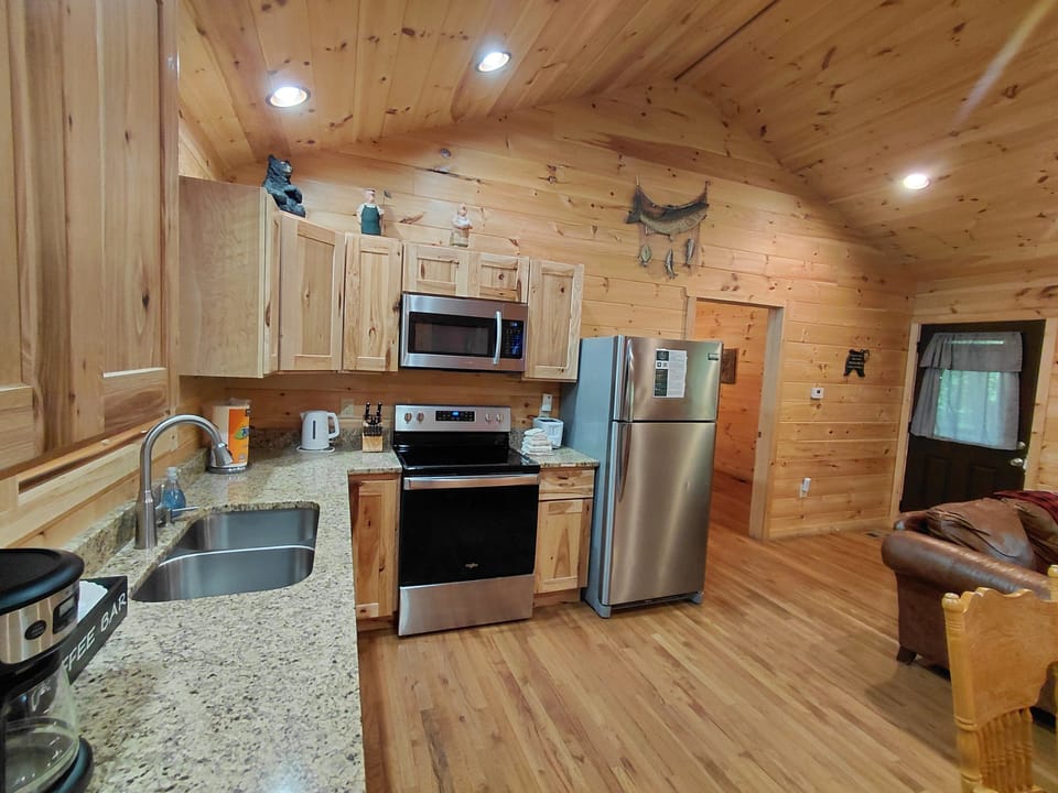 Open-concept cabin interior featuring a full kitchen with stainless steel appliances, granite countertops, wood cabinetry, and a connected living area. Located at Creekside Too near downtown Bryson City in the Smoky Mountains.