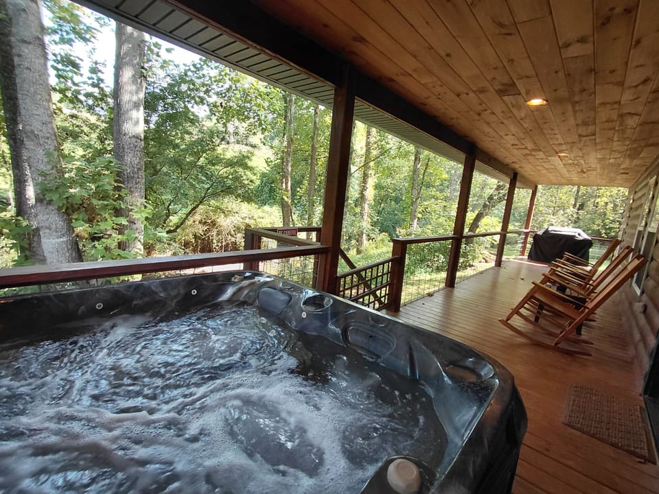Covered deck featuring a bubbling hot tub and rocking chairs with views of the wooded creekside landscape. Outdoor relaxation space at the cabin near Downtown Bryson City.