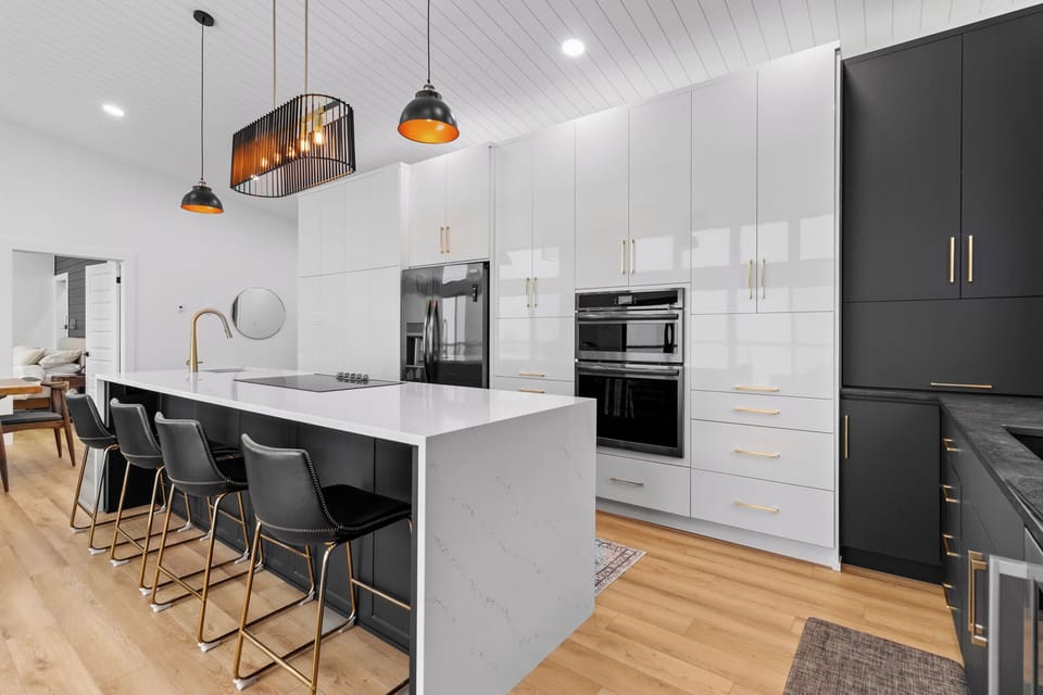 A modern kitchen with a large white island, four black bar stools, white cabinetry, gold handles, a double oven, and a stainless steel refrigerator. Three pendant lights hang above the island, adding a stylish touch.