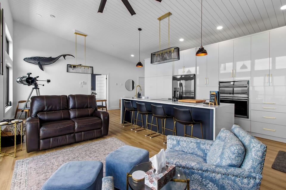 The modern open-concept living area features a sleek kitchen with white cabinetry, black appliances, and a large island with bar seating. There's a comfortable leather sofa, patterned armchairs, and a stylish dining area.