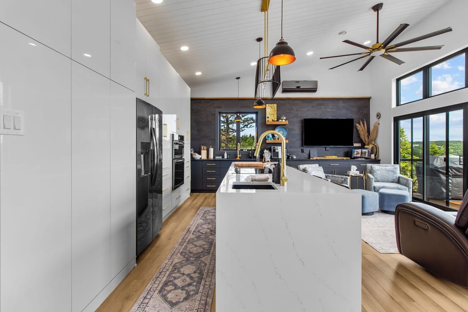 The open-concept kitchen and living area features high ceilings, sleek white cabinetry, a large island with a white marble countertop, black appliances, gold fixtures, and pendant lighting. The living area includes a wall-mounted TV, a large ceiling 
