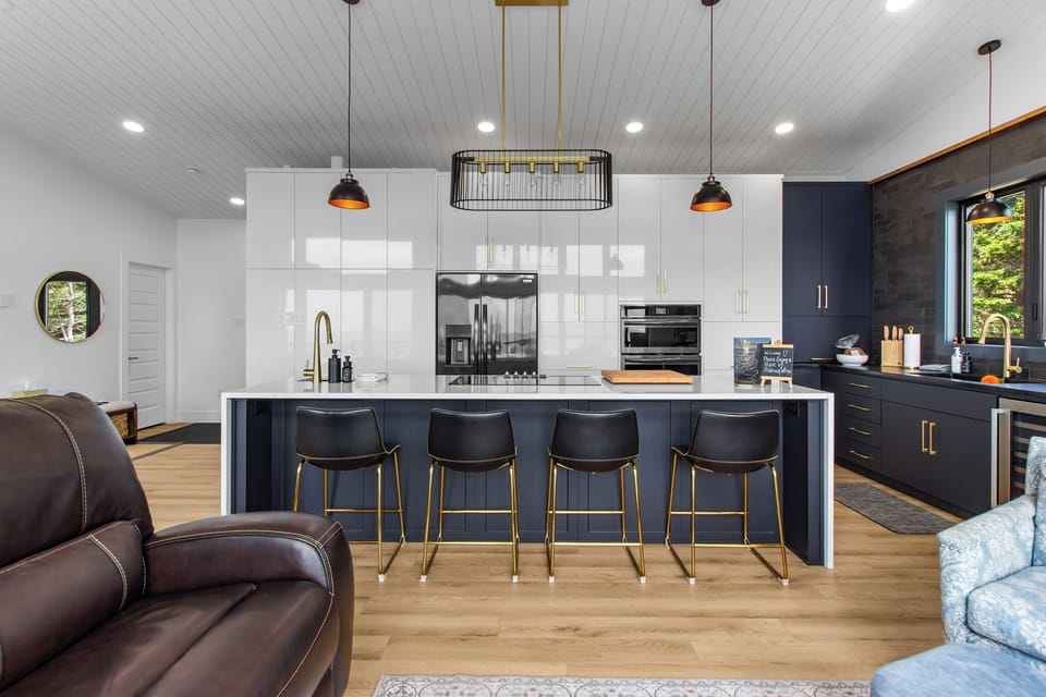 The modern kitchen features a sleek design with a large island, white countertop, black base, four black bar stools, white and black cabinetry, and stainless steel appliances.