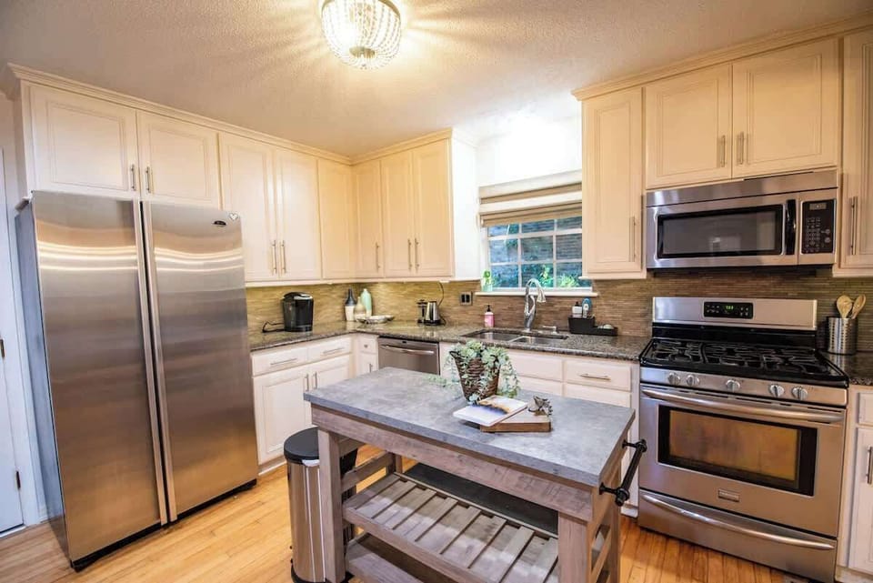 Cook up culinary delights in this spacious, fully equipped kitchen featuring stainless steel appliances—including a large side-by-side refrigerator and gas range—off-white cabinetry, granite countertops, and a central wooden island cart for extra pre