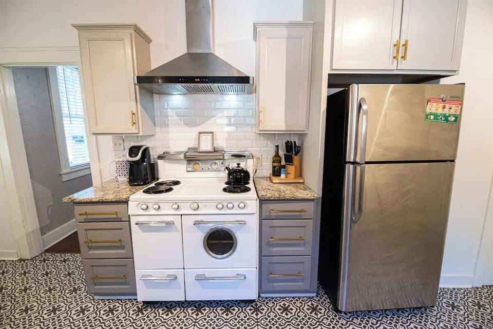 Cook and store meals in this bright kitchen featuring stainless steel appliances, a vintage-style white stove with a range hood, grey cabinetry with brass hardware, and eye-catching patterned tile flooring.