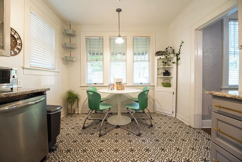 Dine comfortably in this charming breakfast nook featuring a round white pedestal table, teal accent chairs, patterned floor tile, and bright bay windows.