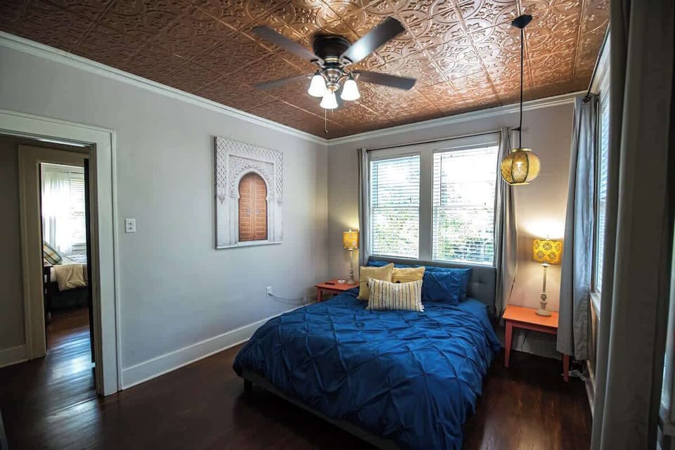 Rest comfortably in this stylish bedroom featuring a rich blue bedding set, matching orange nightstands with warm bedside lamps, polished hardwood floors, and a ornate metallic copper ceiling with a ceiling fan.