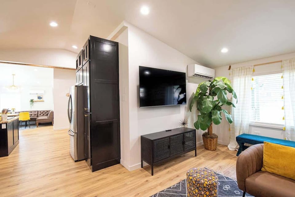Relax in this stylish open-plan living area featuring light wood flooring, a wall-mounted flat-screen TV over a black mesh console, a large potted plant, and a seamless view into the adjacent kitchen and dining space.