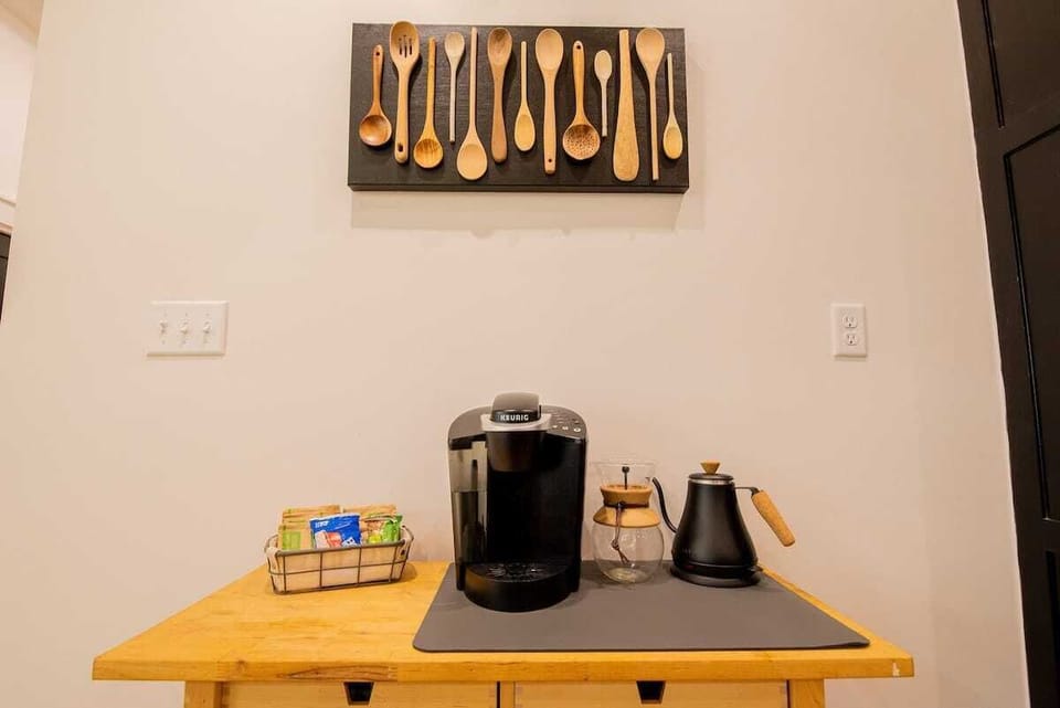 Start your day with a hot drink at this convenient coffee and tea station, equipped with a single-serve coffee machine, french press, black gooseneck kettle, and neatly organized beverage supplies, beneath a decorative wooden spoon wall display.