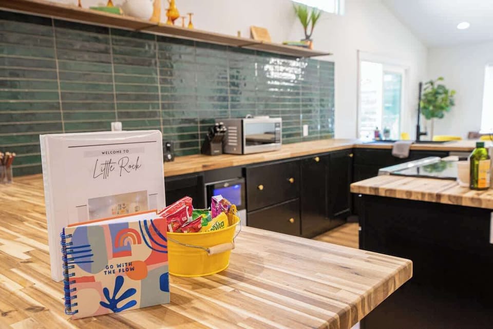 Feel right at home with this welcoming kitchen setup, featuring guest information books and a yellow bucket filled with complimentary snacks on butcher block countertops, surrounded by sleek black cabinets and deep teal subway tiling.