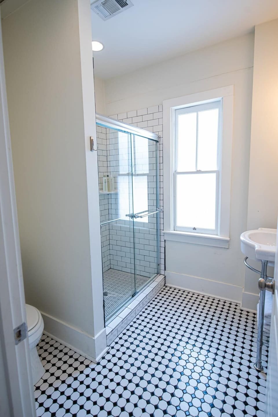 Refresh in this clean bathroom featuring black-and-white patterned mosaic tile floor, a glass-enclosed walk-in shower with white subway tile, a tall window, a pedestal sink, and neutral light walls.
