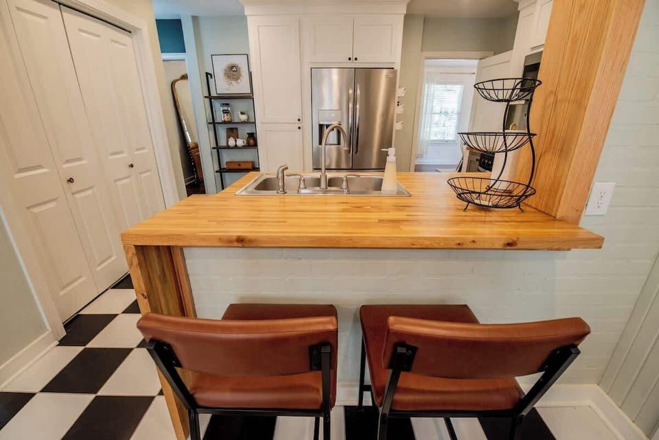 Gather around this inviting breakfast bar, featuring a warm butcher block waterfall countertop paired with brown leather barstools, set over classic black-and-white checkered tile floors with a view into the bright, stainless-steel-equipped kitchen.