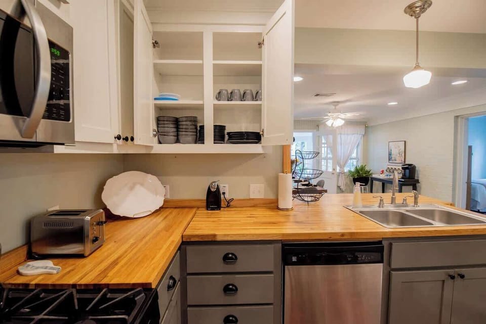 Enjoy full functionality in this well-equipped kitchen, featuring warm butcher block countertops, open upper cabinetry displaying organized dishware, stainless steel appliances including a dishwasher and microwave, and grey lower cabinets.