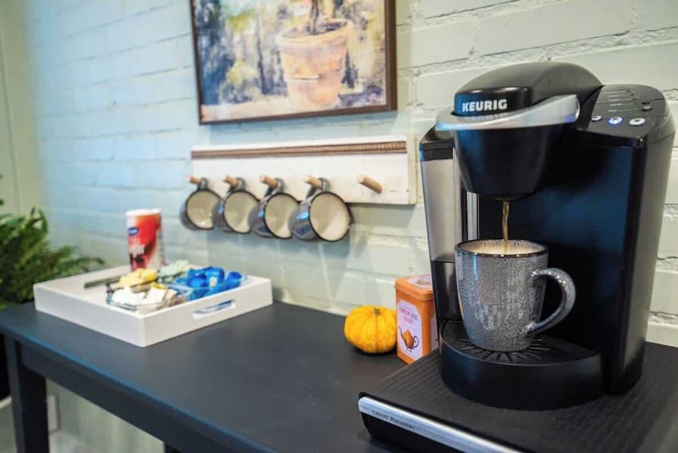 Start your morning right at this cozy coffee bar, equipped with a black Keurig single-serve coffee maker brewing into a grey ceramic mug, a wall-mounted wooden rack holding additional mugs, and a tray stocked with sweet treats and coffee essentials.