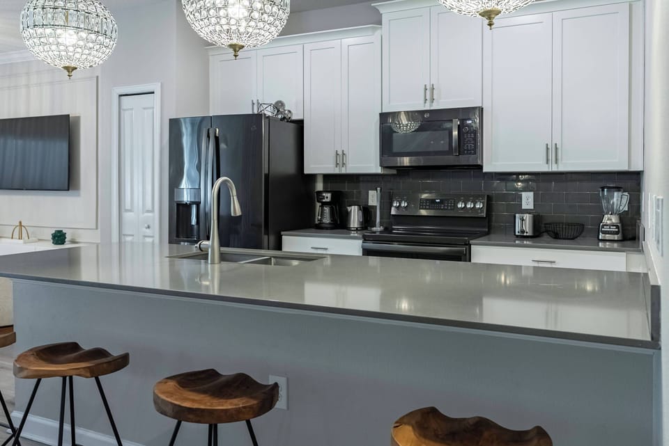 Discover a chic kitchen with sleek gray countertops and white cabinets that beautifully contrast dark accents. Enjoy the warm ambiance created by sparkling pendant lights above the inviting bar seating, ideal for casual meals.