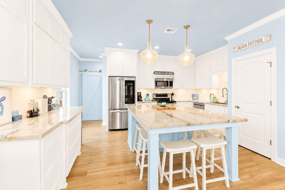 Spacious kitchen featuring stainless steel refrigerator, range, microwave and dishwasher, white cabinets, large island, pendant lights and multiple bar stools.