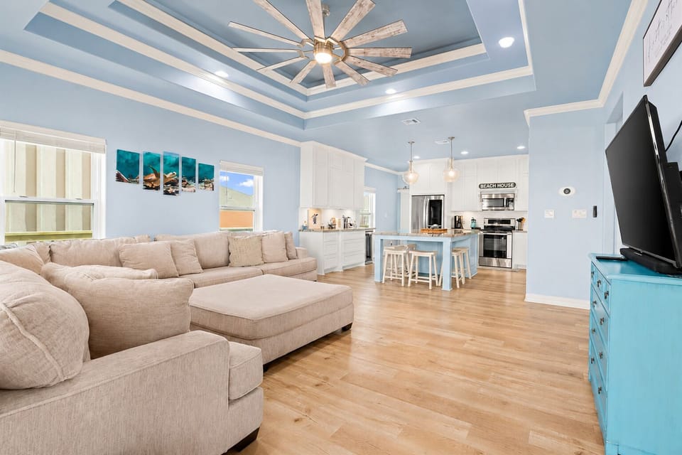 Bright open-concept vacation rental interior featuring a large sectional sofa, soaring tray ceiling, ceiling fan and direct connection to the kitchen and dining area.