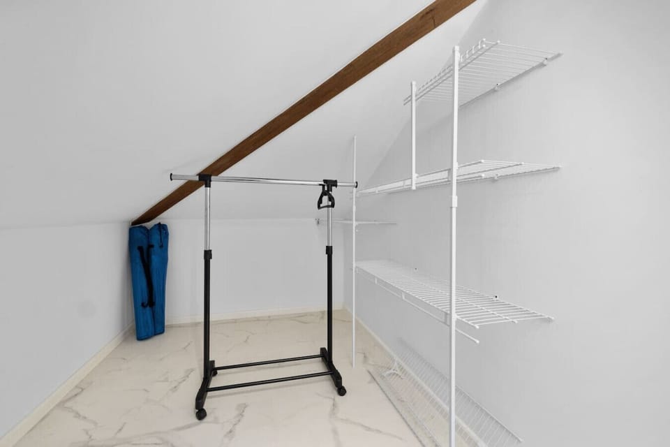 A small attic closet space with a sloped ceiling and a wood beam. A mobile clothing rack stands next to a set of white wire shelves. The floor is white tile with a marble pattern. A blue folded item is visible in the corner.