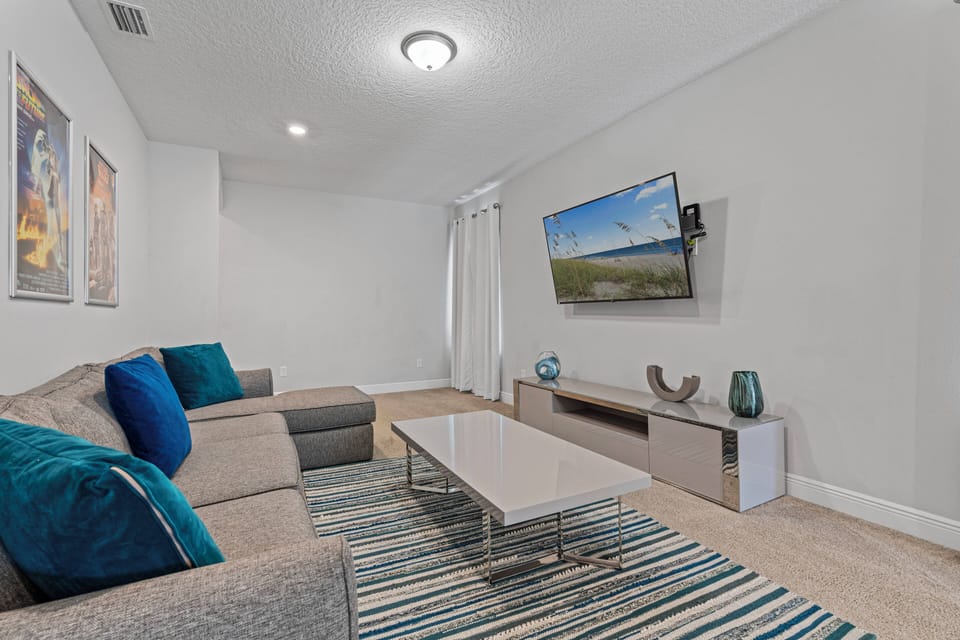Unwind in this stylish living area featuring a cozy sectional sofa, vibrant blue cushions, and a sleek coffee table on a chic striped rug. Sunlight pours through large windows, enhancing the modern decor and inviting you in.