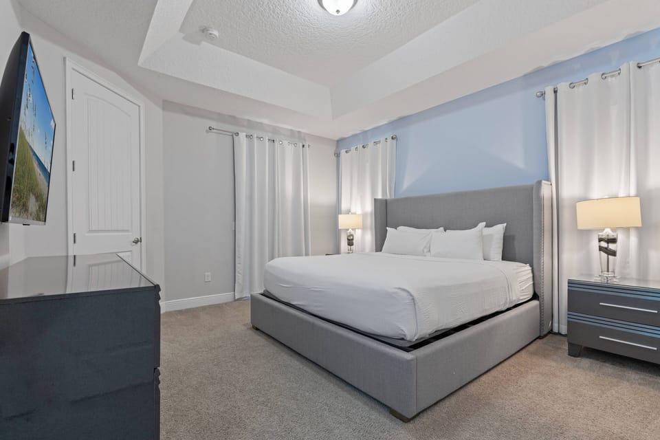 Relax in this serene bedroom featuring a plush king-size bed with crisp white linens. The soft blue walls and elegant curtains create a calming atmosphere, complemented by modern lighting and a flat-screen TV for your convenience.