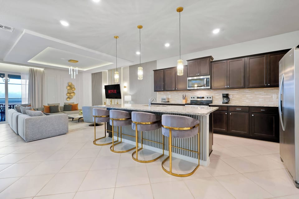 This modern kitchen boasts sleek dark cabinetry and a light countertop, creating a stunning contrast. Enjoy casual meals at the chic island, surrounded by gold-accented bar stools and filled with natural light in the open layout.