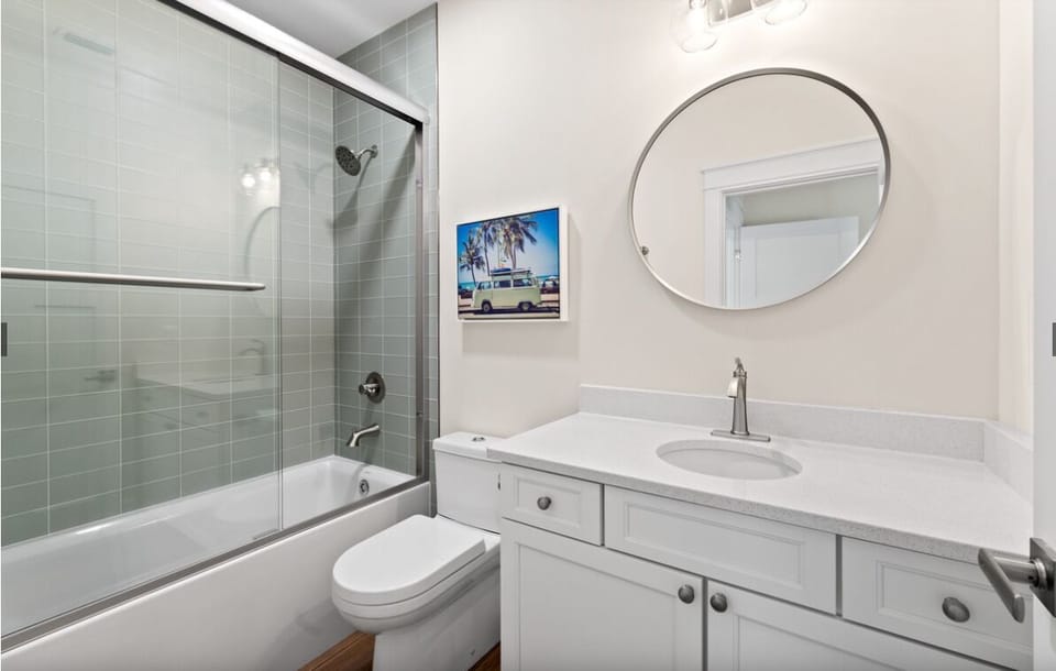 Your pristine bathroom features a sleek glass-enclosed tub-shower combo, elegant white vanity, and charming coastal artwork to brighten your morning routine.