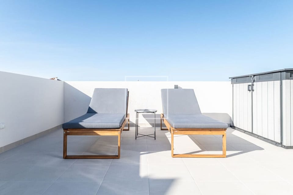 Rooftop terrace features two modern wooden-framed loungers with plush gray cushions and a sleek side table between. Surrounded by white walls under open sky, it’s your serene spot for soaking up the sun in privacy. ☀️🌿