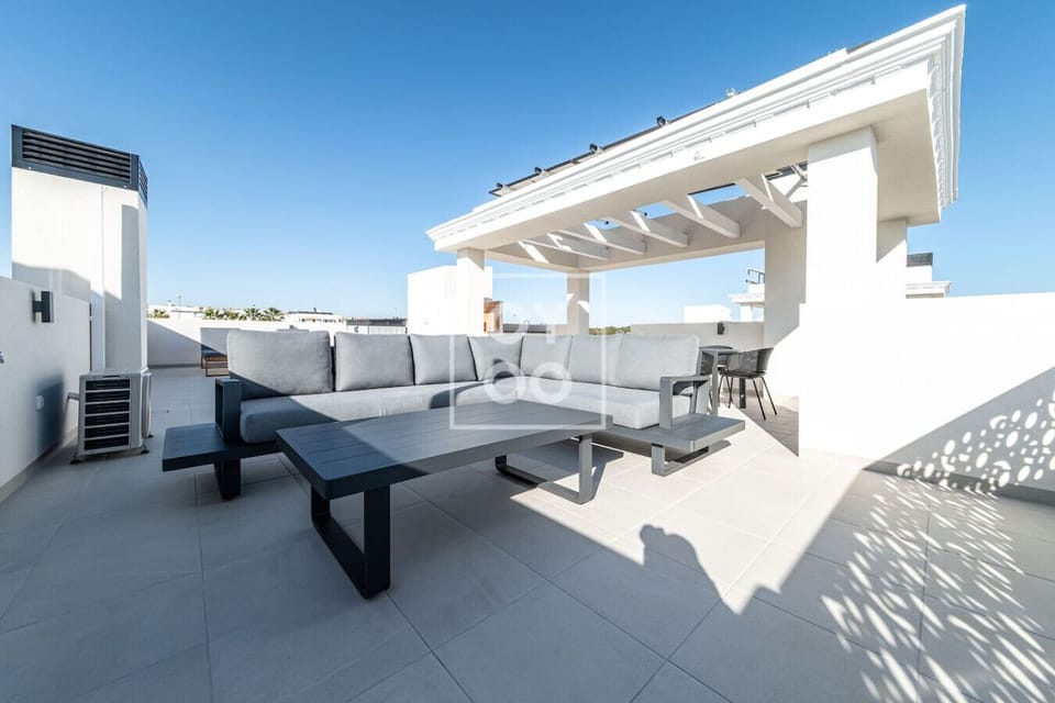 Outdoor rooftop lounge with spacious gray L-shaped sofa and matching coffee table under white beam pergola. Adjacent dining area visible, all framed by clear blue sky for stylish al fresco relaxation. 🛋️🍹☀️