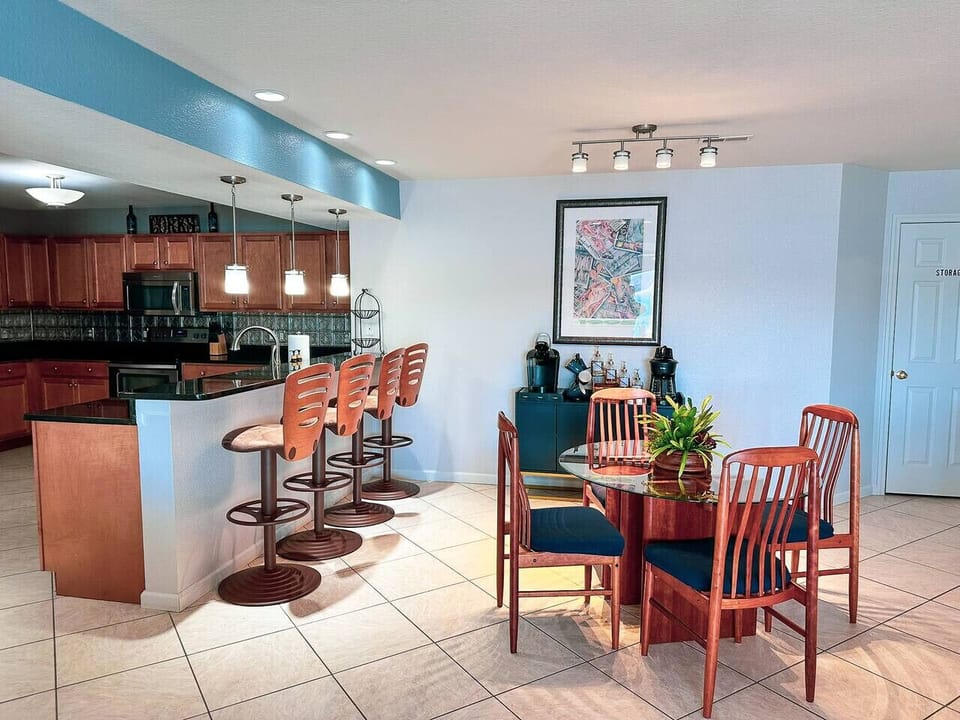 A spacious, open-concept kitchen and dining area featuring a sleek glass-top dining table, stylish pendant lighting, and a fully stocked coffee and beverage station.