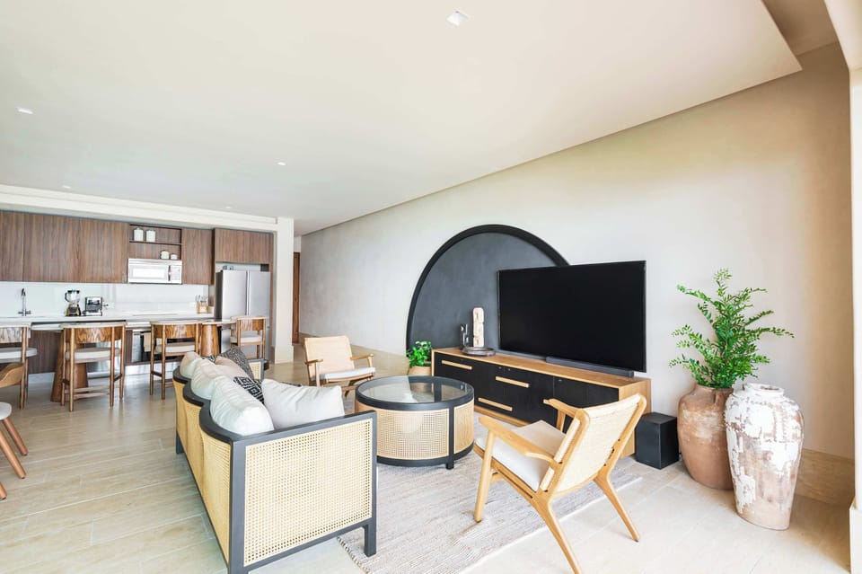 Two light-colored armchairs with woven backs flank the console, positioned on either side of a round, glass-topped coffee table with a woven base. A large, comfortable sofa with light-colored cushions and a decorative, patterned base faces the TV. 
