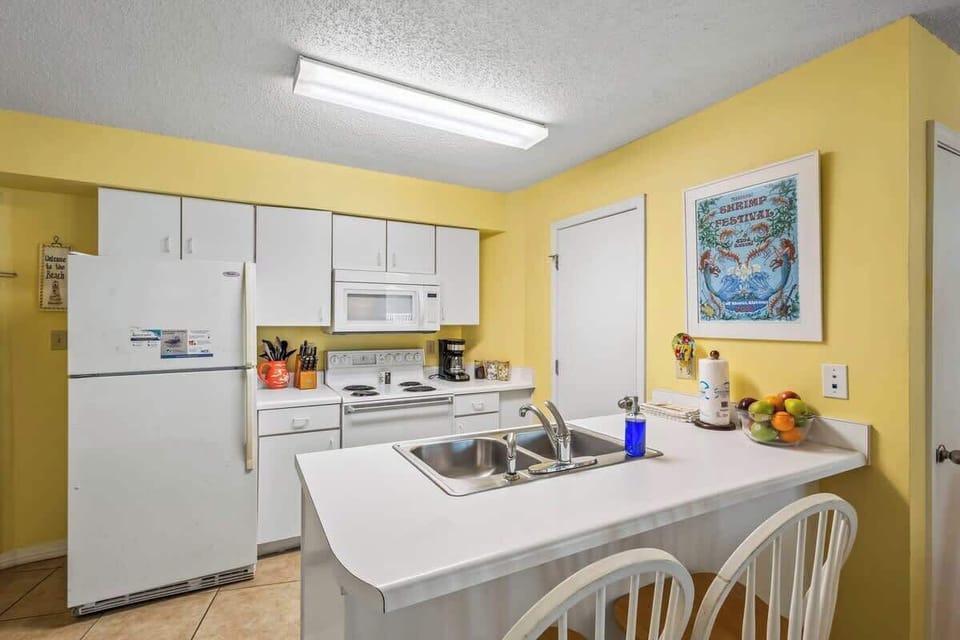 This well-lit kitchen features white cabinetry, a full-size refrigerator, a microwave, and an electric stove. The spacious countertop with a double sink makes meal prep a breeze. Perfect for home-cooked meals on your beach getaway!