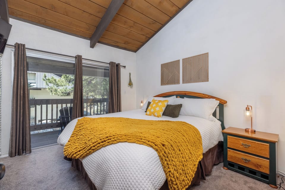 Retreat to the master bedroom, where vaulted wood-beam ceilings, plush bedding, and a cozy golden throw create a warm and inviting sanctuary. Wake up to tranquil views through sliding glass doors that open onto a private balcony, blending comfort and mountain charm for an unforgettable stay.