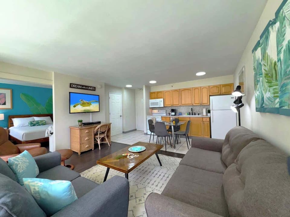 A comfy, open-concept apartment featuring a spacious living room with a pull-out sleeper sofa, a couch, a full kitchen with wood cabinets, and a peek into a bright, tropical-themed bedroom