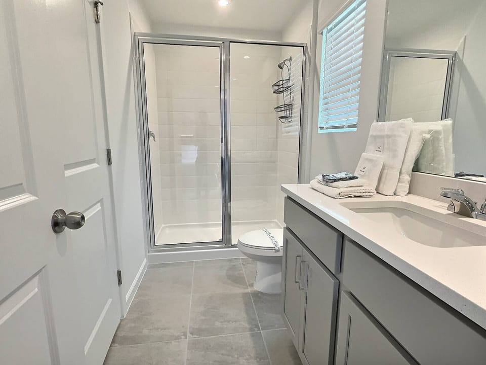 Bright and airy, this bathroom features sleek gray tile flooring and a spacious glass shower that invites relaxation. The contemporary vanity, adorned with fresh towels, adds a touch of modern comfort to your stay.