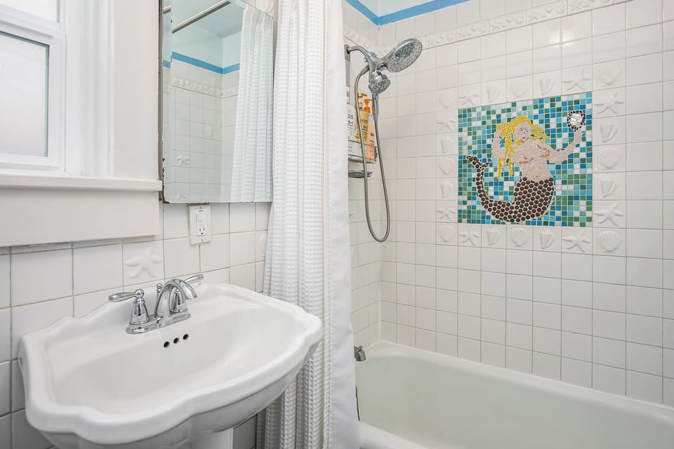 Starfish-embossed white tile and a handcrafted mermaid mosaic make the tub-shower combo genuinely fun — the dual-function showerhead with handheld wand and provided shampoo and for safety a grab bar is next to the shower handle.
