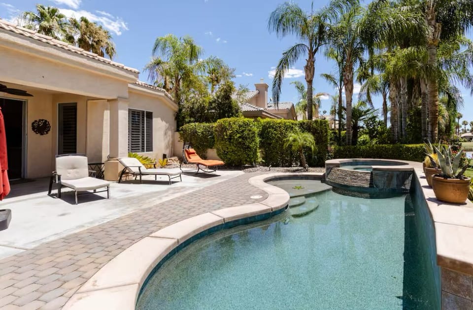 Sun-soaked patio with pool, spa & lounge chairs framed by swaying palms