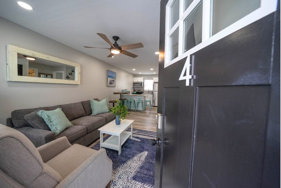 Open-concept living area with comfortable sofa, modern decor, ceiling fan, and fully equipped kitchen with bar seating. Bright and welcoming space, perfect for relaxing, dining, and enjoying a coastal stay.