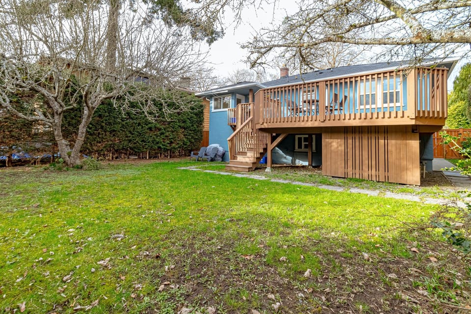 Grassy backyard with mature trees and a raised wooden deck featuring outdoor seating and stairs.